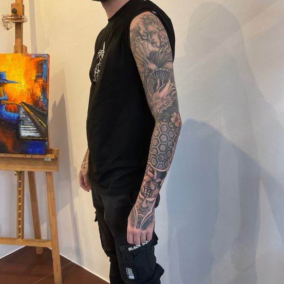 Ein Mann mit einem vollständigen Arm-Tattoo steht neben einem Staffelei mit einem Gemälde. Er trägt ein schwarzes Shirt und Hose.