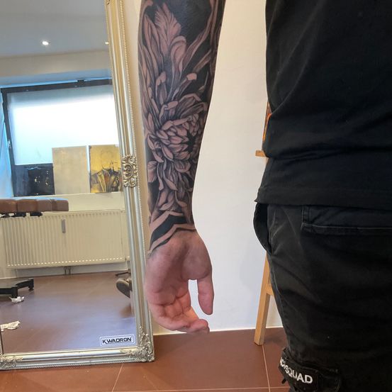 Eine Person mit einem schwarzen Shirt und Hose steht vor einem Spiegel und zeigt ein Tattoo am Arm.