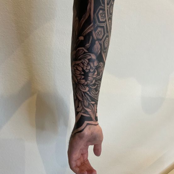 Ein Nahaufnahme eines linken Unterarms, der ein detailliertes Tattoo mit einer großen Blume und einem geometrischen Muster zeigt. Das Tattoo ist vor einem schlichten weißen Hintergrund zu sehen.