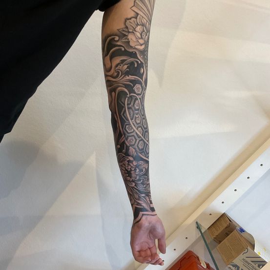 Eine Nahaufnahme eines Armes mit einem Ärmeltattoo. Das Tattoo zeigt ein Blumendesign und ein sechseckiges Muster.