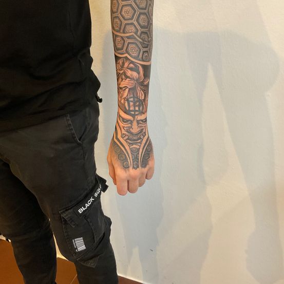 Eine Person mit einem Tattoo eines Gesichts, Sechsecke und Blumen am Arm steht vor einer weißen Wand.