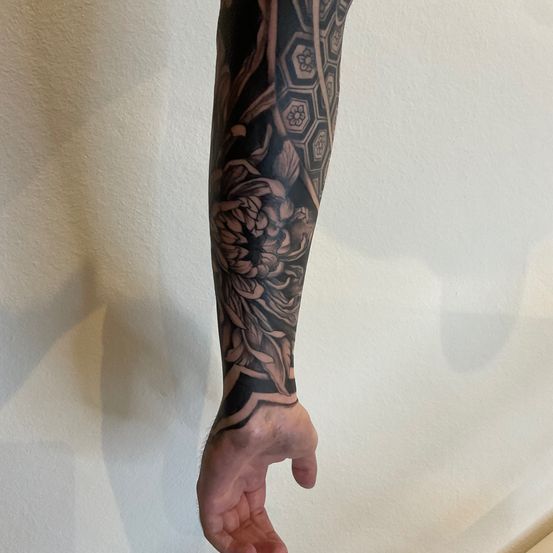 Eine Nahaufnahme eines Unterarms mit einem großen, detaillierten Tattoo, das eine Blume und ein sechseckiges Muster zeigt. Das Tattoo ist in Schwarz- und Grautönen gehalten.