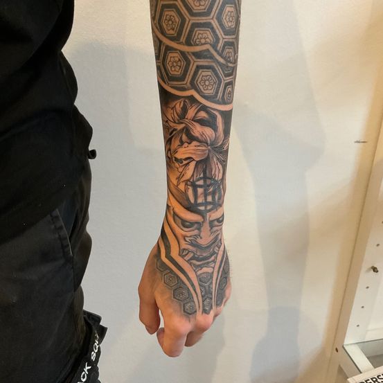 Eine Person zeigt ein Unterarm-Tattoo mit einem stilisierten Masken- und Blumendesign, wobei geometrische Muster den Arm entlang verlaufen.
