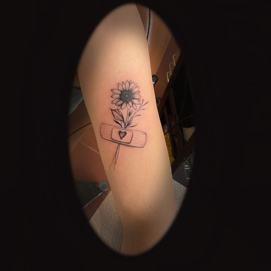 Ein Tattoo am Arm einer Person zeigt eine Sonnenblume mit Blättern, begleitet von einem Verband mit Herzsymbol.