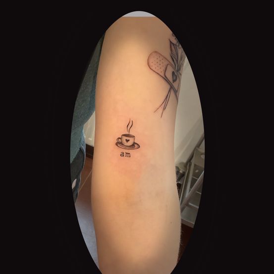 Eine Tätowierung am Arm einer Person zeigt eine Kaffeetasse mit Dampf, ein Herz und einen Verband. Das Wort 'am' ist ebenfalls darunter tätowiert.
