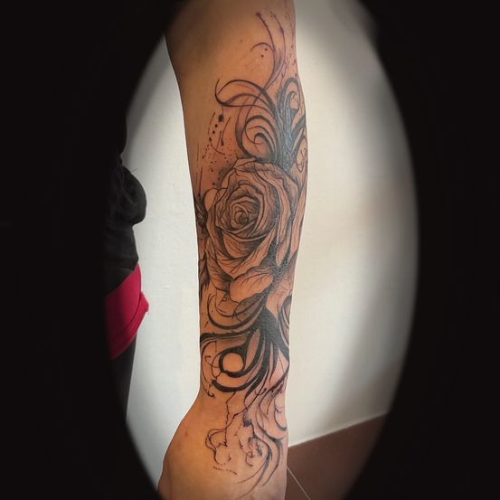Der Arm einer Person zeigt ein detailliertes schwarzes und graues Tattoo einer Rose mit Wirbeln und abstrakten Mustern, die sich von den Blütenblättern aus erstrecken.