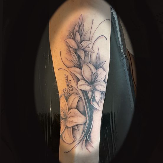 Ein detailliertes Tattoo von weißen Lilien mit grünen Blättern am Arm einer Person. Die Blüten sind schattiert, um einen dreidimensionalen Effekt zu erzeugen.