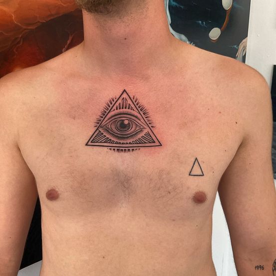 Die Brust eines Mannes zeigt zwei Tattoos. Eines ist ein Auge in einem Dreieck und das andere ist ein Dreieck darunter. Die Brust ist haarlos, mit einem kleinen Höcker auf der linken Seite.