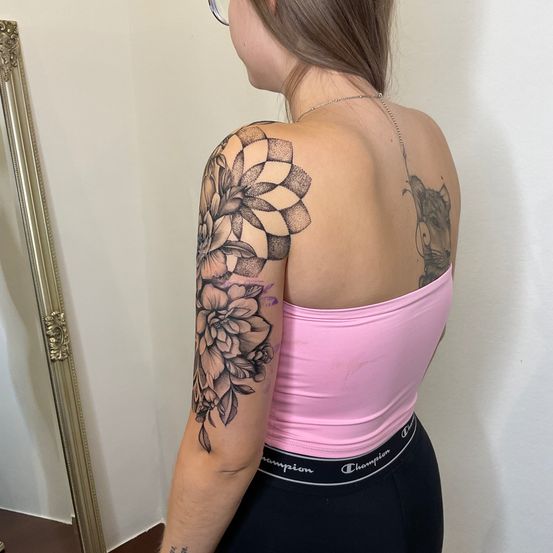 Eine Frau mit Schultertattoos, eines eine Blume und das andere eine Katze, steht vor einem Spiegel und trägt ein rosa Top und schwarze Hose.