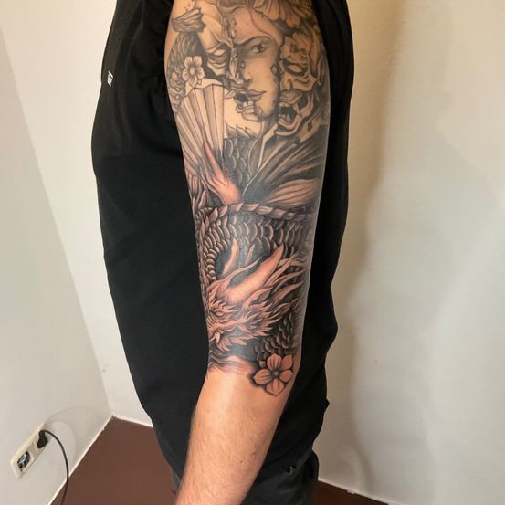Eine Person in einem schwarzen T-Shirt zeigt eine Tätowierung am linken Arm mit einem Drachen, einer Maske und Blumen.