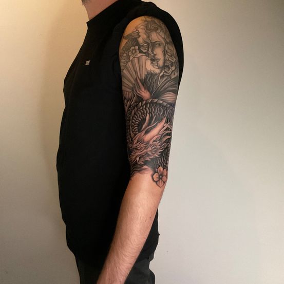 Ein Mann in einem schwarzen Shirt steht gegen eine weiße Wand und zeigt ein detailliertes Tattoo auf seinem Arm, das einen Drachen und Blumendesigns darstellt.