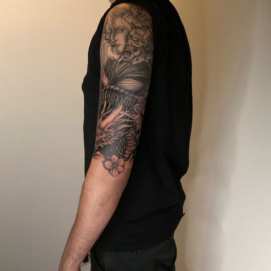 Eine Person steht mit dem Rücken zur Seite. Sie hat ein detailliertes Tattoo auf dem linken Arm, das einen Drachen und das Gesicht einer Frau zeigt.