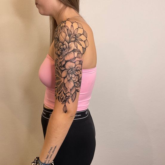 Eine Frau mit einem Blumentattoo am Arm, das in einem rosa Tanktop und schwarzen Leggings trägt, steht vor einer weißen Wand.