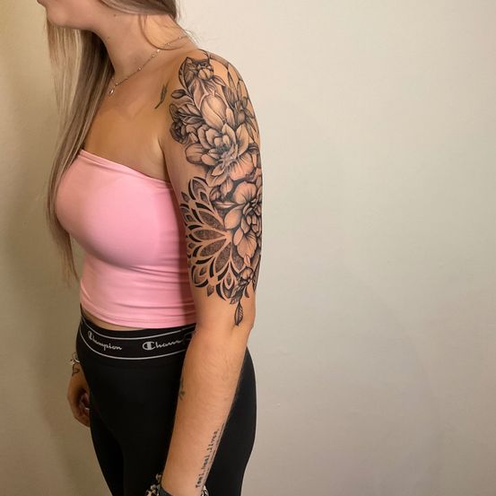 Eine Frau mit langen blonden Haaren steht und zeigt ihren linken Arm mit einem Blumentattoo. Sie trägt ein rosa Top und schwarze Leggings.