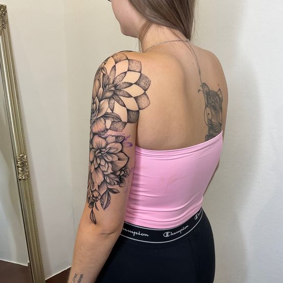 Eine Frau mit einem großen Blumentattoo auf dem linken Arm und einem Katzentattoo auf der rechten Schulter, die vor einem Spiegel steht.