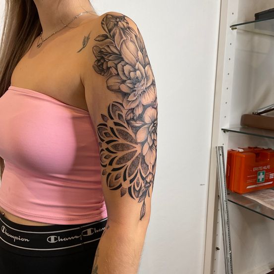Eine Frau mit einem rosa Oberteil und schwarzen Hosen zeigt ein großes Tattoo auf ihrem rechten Arm. Das Tattoo ist ein detailliertes Blumendesign mit Blütenblättern, die sich bis zur Schulter erstrecken.