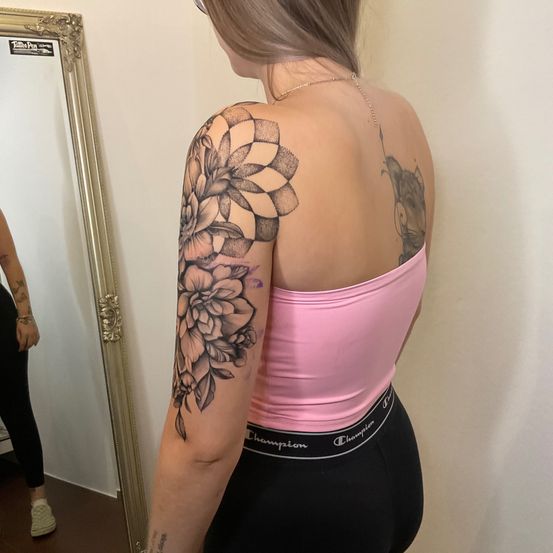 Eine Frau mit einem Blumentattoo am Arm, die sich selbst im Spiegel betrachtet, trägt ein rosa Oberteil und schwarze Leggings.