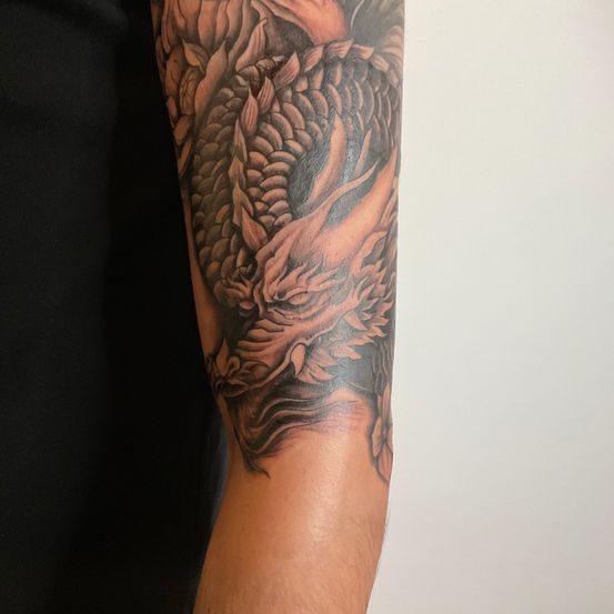 Ein detailliertes Drachen-Tattoo befindet sich am rechten Unterarm einer Person. Der Drache ist mit feurigen Flammen und komplizierten Mustern dargestellt.