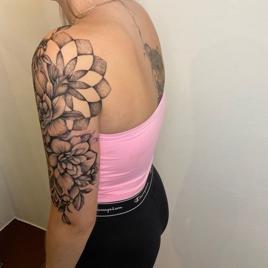 Eine Frau mit einem Blumentattoo auf dem Schulter steht vor einer weißen Wand und trägt ein rosa Tanktop und schwarze Leggings.