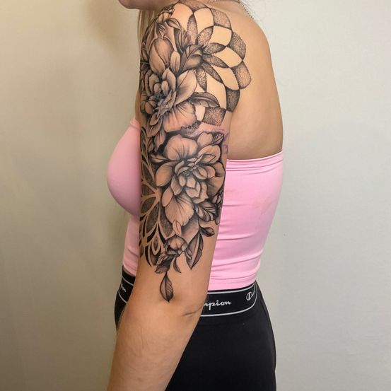 Eine Frau mit einer Tätowierung von Blumen am Oberarm steht in einem Raum. Sie trägt ein rosa Tanktop und schwarze Leggings.