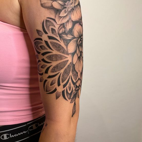 Eine Frau mit einem rosa Top zeigt ein detailliertes Blumentattoo am linken Arm. Das Tattoo ist schwarz und grau mit einem geometrischen Design.