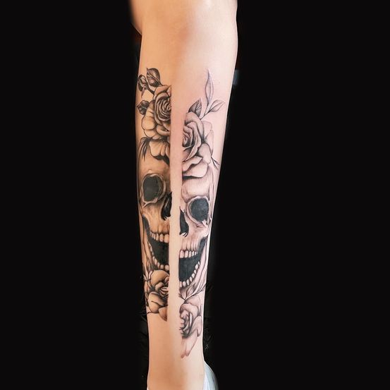 Ein Tattoo von zwei Schädeln mit Blumen auf dem Arm einer Person, in der Mitte durch eine vertikale Linie geteilt.