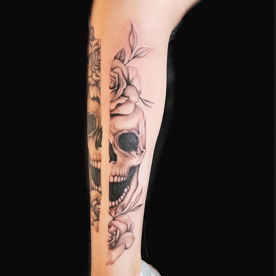 Eine Tätowierung von zwei Schädeln und Rosen am Arm einer Person. Die Schädel sind vertikal geteilt und mit Blumen verziert.