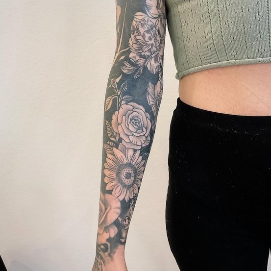 Eine Person mit einem Armtattoo verschiedener Blumen, einschließlich Rosen und Sonnenblumen, vor einer weißen Wand.