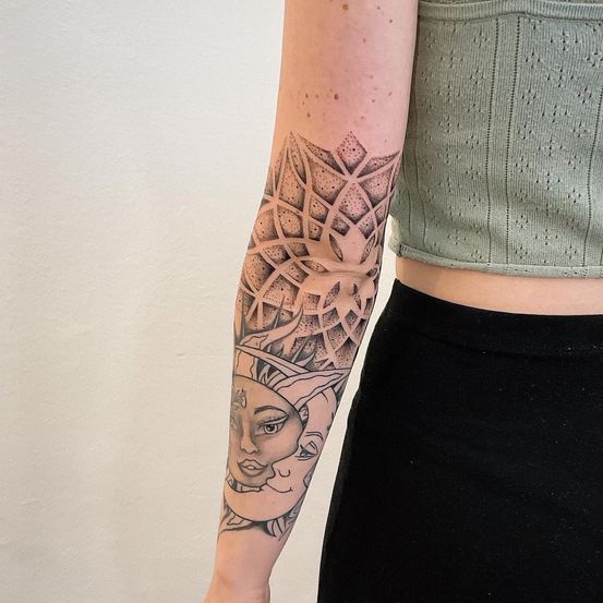 Der Unterarm einer Person zeigt ein Tattoo von Sonne und Mond, das geometrische Muster und Gesichtszüge vereint.