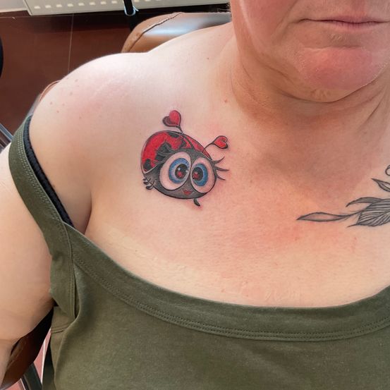 Eine ältere Frau, die ein grünes Tanktop trägt, hat eine zeichentrickartige Tätowierung einer Marienkäfer auf ihrer Schulter.