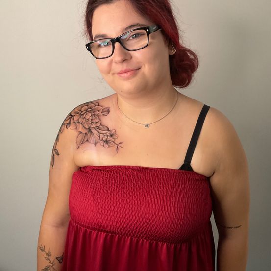 Eine Frau mit roten Haaren und einer Brille posiert für ein Foto. Sie hat ein Blumentattoo auf der Schulter und trägt ein rotes ärmelloses Top und eine Halskette.