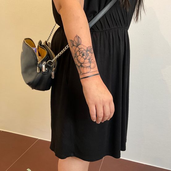 Eine Frau in einem schwarzen Kleid, die eine schwarze Handtasche hält, hat ein Blumentattoo auf ihrem linken Unterarm.