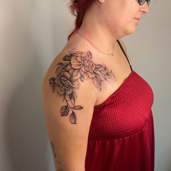 Eine Frau mit roten Haaren trägt ein rotes Kleid und eine Halskette. Sie hat ein Blumentattoo auf der Schulter.