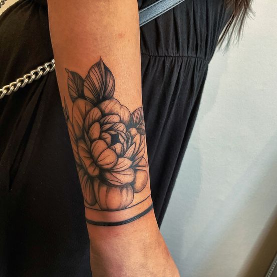 Ein Nahaufnahme des Arms einer Frau zeigt ein detailliertes Blumentattoo, einschließlich Blütenblätter und Blätter, auf ihrem Unterarm.