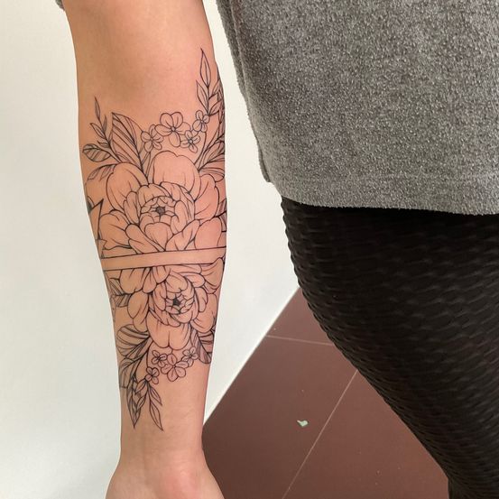 Eine Person mit einem Blumentattoo am Arm, bestehend aus einer großen Blume mit Blättern und kleineren Blumen, vor weißem Hintergrund.