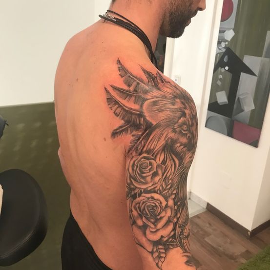 Ein Mann mit einer Tätowierung eines Vogels und Rosen auf seinem linken Arm, steht in einem Raum.