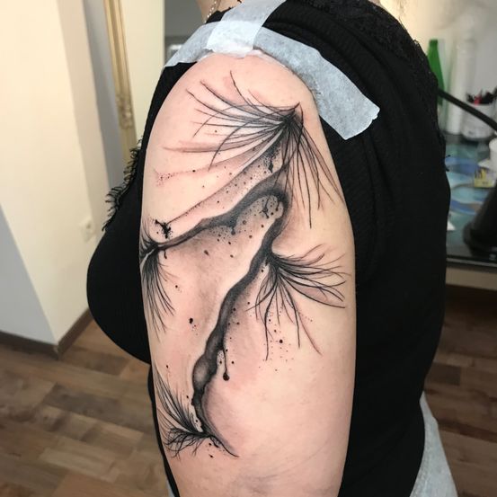 Eine Person mit einem schwarzen Tintentattoo eines federartigen Designs am linken Arm, das in einem Raum steht.