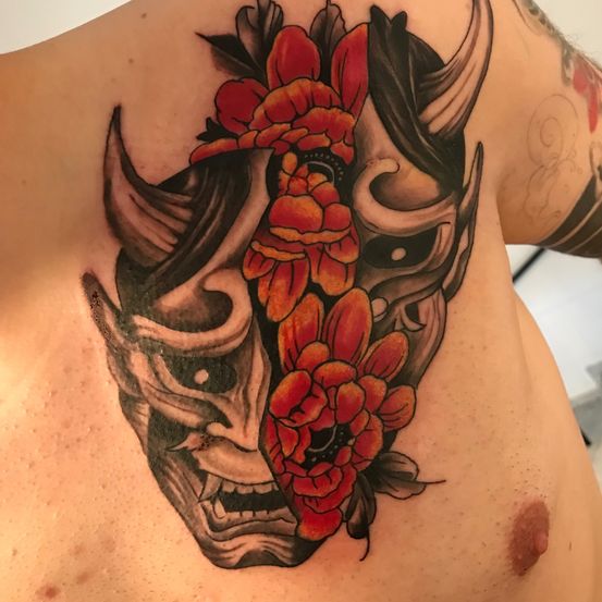 Ein Tattoo eines japanischen Dämonenmaskes mit roten Blumen auf der Schulter. Die Maske ist in zwei Hälften geteilt.