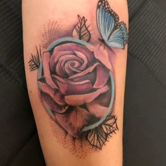 Ein detailliertes Tattoo am Arm zeigt eine Rose in einem runden Rahmen, umgeben von zwei Schmetterlingen und Blättern.