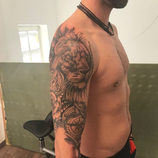 Ein bärtiger Mann ohne Oberteil mit einem Löwen-Tattoo auf dem linken Arm, das einen Kompass und eine Rose zeigt. Er steht vor einer grünen Wand.