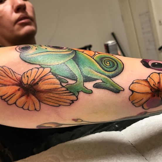 Ein Nahaufnahme des Armes einer Person zeigt eine farbenfrohe Tätowierung eines Chamäleons, das auf einer orangefarbenen Blume sitzt. Das Chamäleon ist detailliert mit grünen und orangefarbenen Farbtönen und die Blume ist lebhaft mit einer Mischung aus orangen und gelben Blütenblättern.