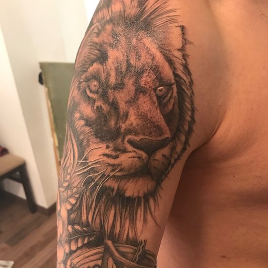 Eine Nahaufnahme des Arms eines Mannes mit einem detaillierten Löwen-Tattoo mit fiesen Augen, einem fließenden Mähnen und einem wilden Ausdruck. Der Hintergrund besteht aus einer weißen Wand und einem Holzboden.