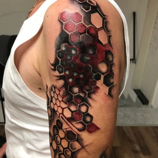Ein Mann mit einem weissen ärmellosen Oberteil zeigt eine Tätowierung an seinem Arm mit einem geometrischen Muster aus Sechsecken in Schwarz-, Rot- und Grautönen.