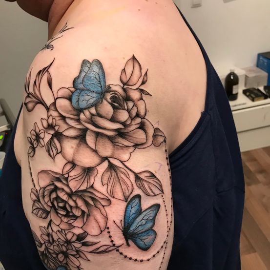 Eine Person zeigt ein Tattoo auf ihrer Schulter, das einen Blumenstrauß und zwei blaue Schmetterlinge zeigt. Das Tattoo ist detailliert und farbenfroh.