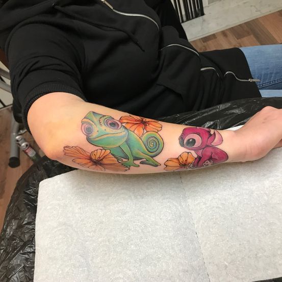 Eine Person in einem schwarzen Jacken sitzt mit einer bunten Tätowierung am Arm, die einen Chamäleon und eine rosa Kreatur zeigt. Die Tätowierung umfasst Blumen und befindet sich am Unterarm.