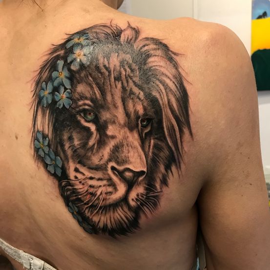 Der Rücken einer Person zeigt ein detailliertes Tattoo eines Löwen mit blauen Blumen um seine Mähne. Das Tattoo ist lebhaft farbige und sorgfältig gestaltet.