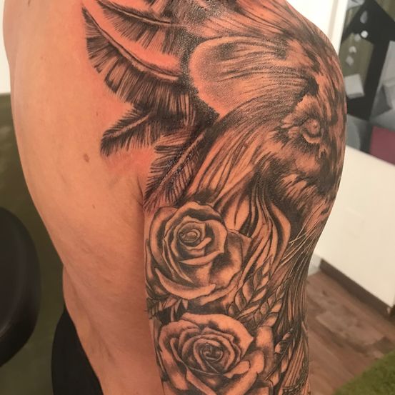 Ein detailliertes Tattoo am Oberarm einer Person, das einen Vogel mit Flügeln und zwei Rosen darunter zeigt. Der Vogel und die Rosen sind in einem Schwarz-Rot-Farbschema gehalten.