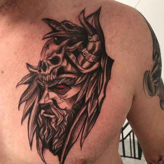 Eine Tätowierung eines bärtigen Mannes mit roten Augen, einem Schädel und einem Raben auf dem Kopf befindet sich auf der Brust einer Person.