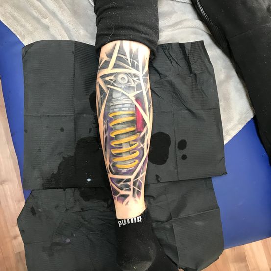 Ein Nahaufnahme eines Unterarms mit einem mechanischen Tattoo. Das Tattoo ist detailliert und umfasst eine Spiralfeder und andere Maschinenteile.