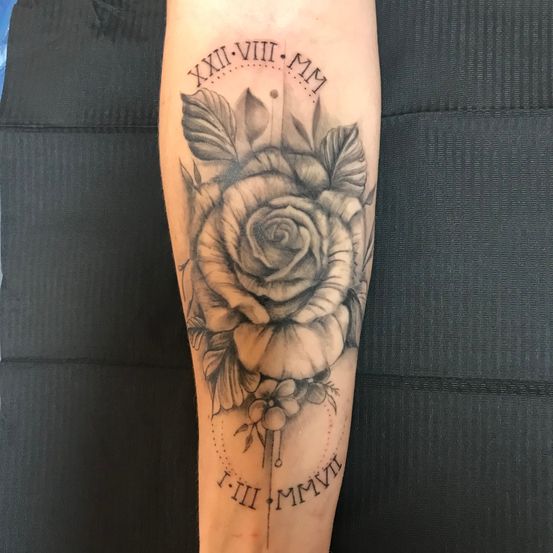 Ein Tattoo am Arm einer Person zeigt eine Rose mit Blättern und einem Stiel. Die Rose ist von römischen Ziffern umgeben. Das Design enthält auch einen kleinen Punkt über der Rose.